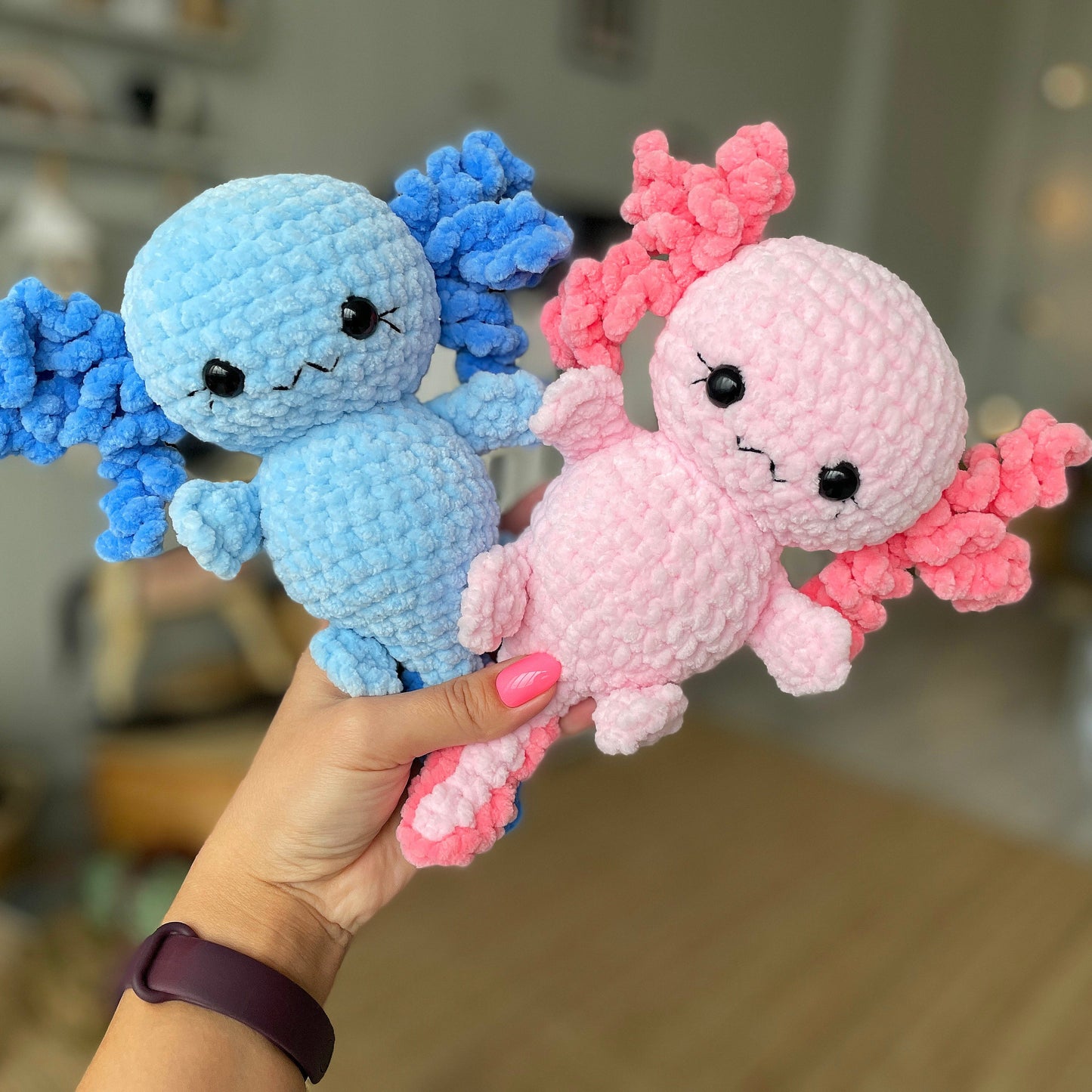 Two small crochet dolls, one blue and one pink, held in a hand with a blurred indoor background.